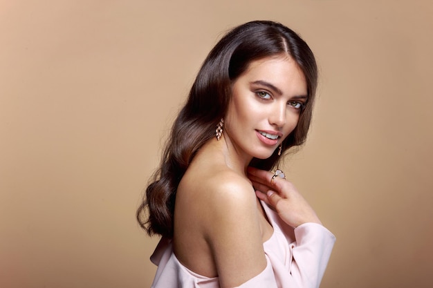 Young woman dressed in a pink blouse with bare shoulder, brown wavy hair, model with bracelet and earrings over brown background.