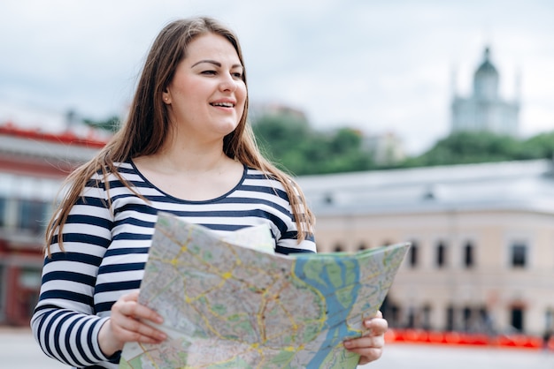 Giovane donna vestita con un maglione a righe, con una mappa in mano, all'aperto, cammina per le strade della città