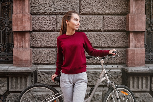 壁の近くに立って街を自転車で歩くセーターを着た若い女性