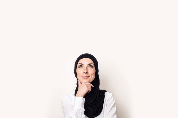 Giovane donna vestita con una camicia bianca e hijab si tiene la mano al mento e alza lo sguardo con una faccia pensierosa