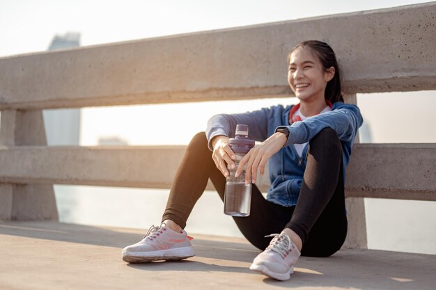 若い女性は、市内で朝のトレーニングをジョギングした後、水を飲み、座って休憩します。首都で健康に暮らす街。運動、フィットネス、ジョギング、ランニング、ライフスタイル、健康的なコンセプト。