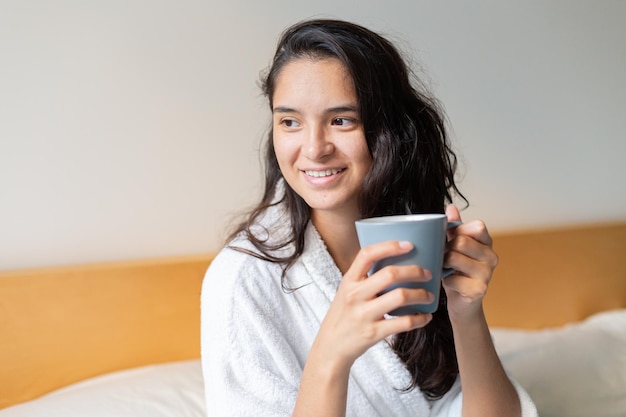 バスローブ姿でベッドでコーヒーを飲む若い女性