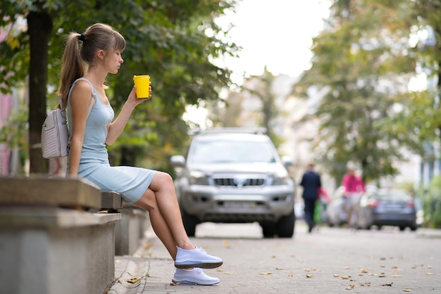 サマーパークの街のベンチに座っている紙コップからコーヒーを飲む若い女性。