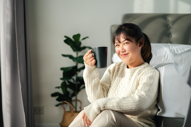Фото Молодая женщина пьет кофе в спальне, держа кофейную чашку двумя руками.