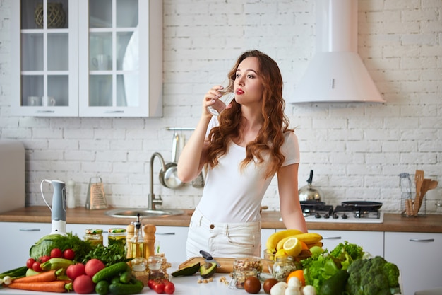 若い女性が台所でガラスから新鮮な水を飲みます。健康的なライフスタイルと食事。健康、美容、ダイエットのコンセプトです。