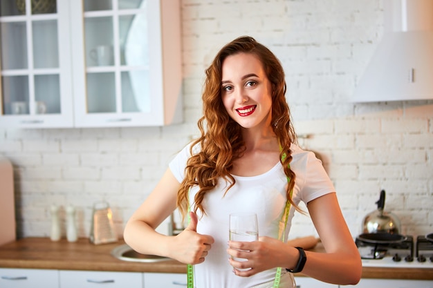 Giovane donna che beve acqua dolce da vetro e che mostra i pollici su nella cucina. stile di vita sano e mangiare. salute, bellezza, concetto di dieta.