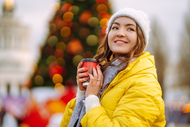 新年の木の近くを歩きながらホットコーヒーを飲む若い女性。