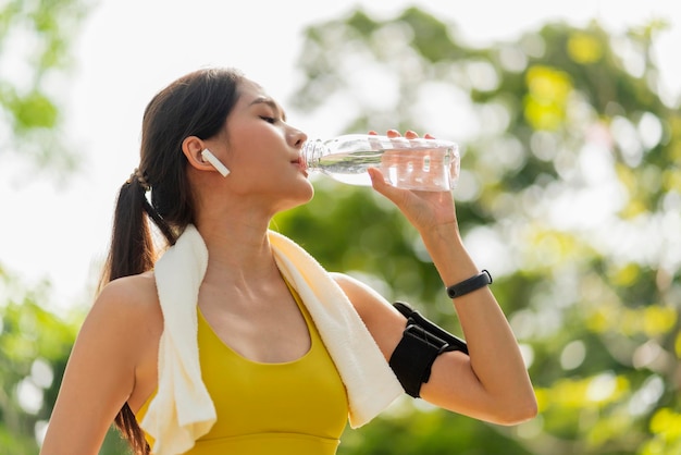 若い女性がボトルから水を飲む運動やスポーツの後のアジアの女性の飲料水日没の夜に運動した後、帽子をかぶった飲料水を身に着けている美しいフィットネスアスリートの女性