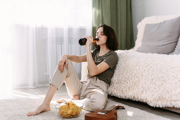 A young woman drinks beer and eats chips at home