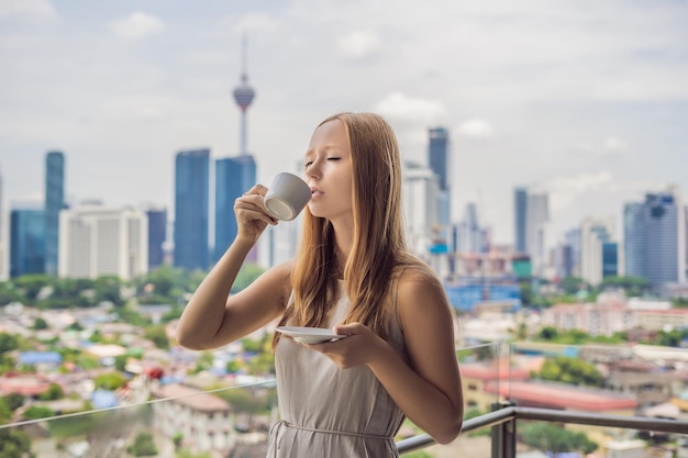 若い女性は、大都市と高層ビルを見下ろすバルコニーで朝コーヒーを飲みます