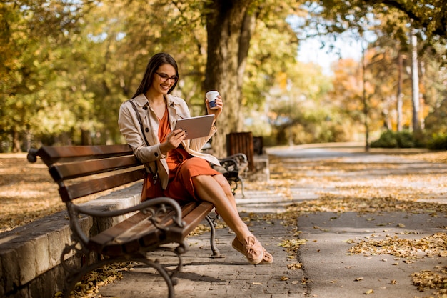 若い女性がコーヒーを飲み、公園でデジタルタブレットを使用する