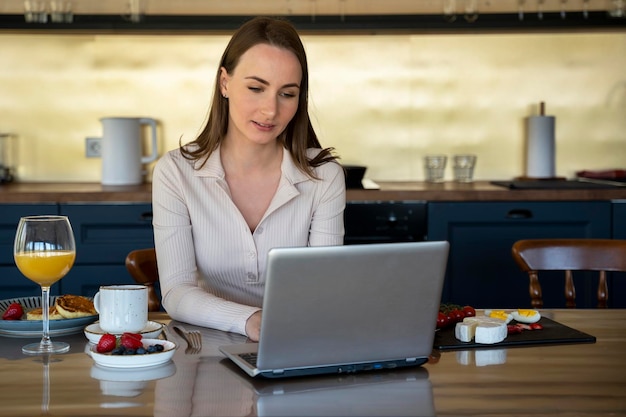 Young woman drinks orange juice and works on her laptop at breakfast at home