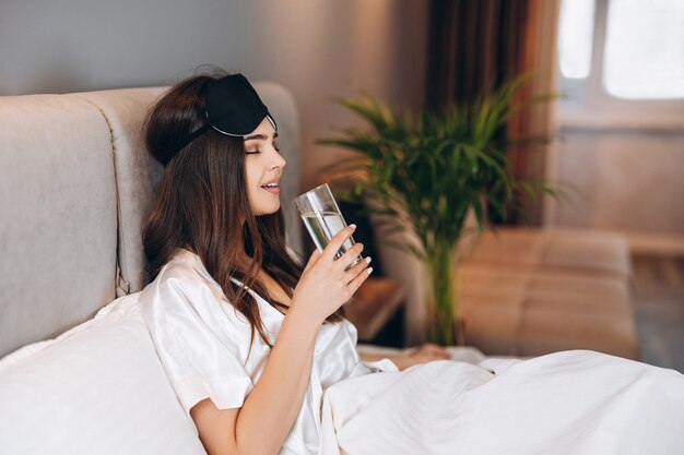 Photo young woman drinks water in the bed. model with brown hair with a glass of water in the bed in the bedroom. drink a glass of water in the morning or before bed. young woman enjoying drinking water.