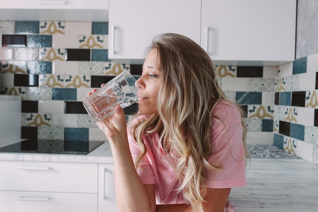 Photo a young woman drinks water from a large transparent glass standing in the kitchen