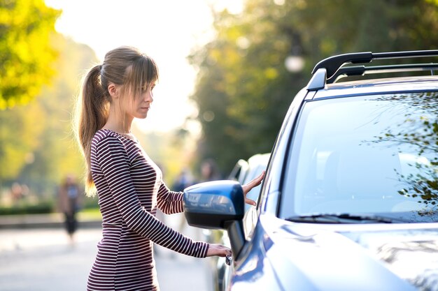 街の通りで彼女の車の横に立って暖かい夏の日を楽しんでいる若い女性の運転手。旅行と休暇のコンセプト。