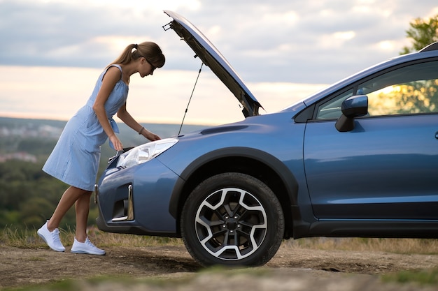 Young woman driver standing near a broken car with open hood having trouble with her vehicle