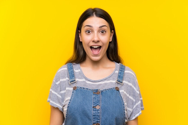 Young woman in dungarees isolated on yellow  with surprise facial expression