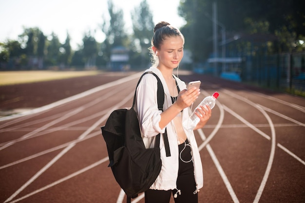 Young woman in earphones using cellphone with bottle of water in hand while spending time on racetrack of stadium