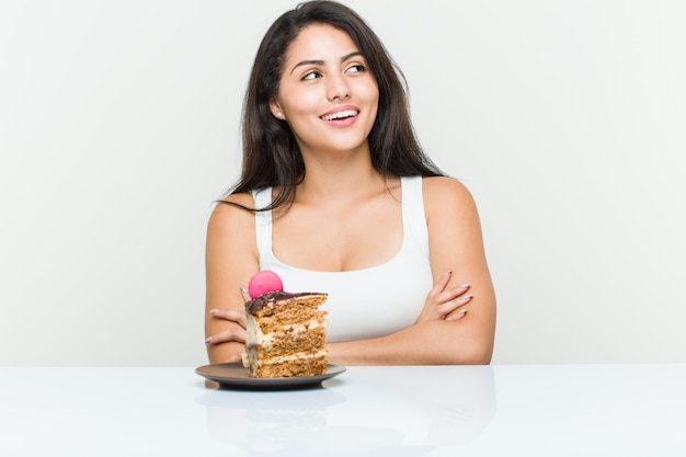 Young woman eating a cake smiling confident with crossed arms.