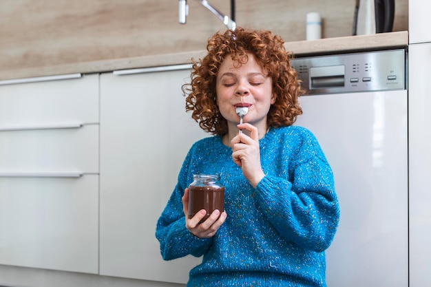 young woman eating chocolate from a jar while sitting on the wooden kitchen floor Cute ginger girl indulging cheeky face eating chocolate spread from jar using spoon savoring every mouthful