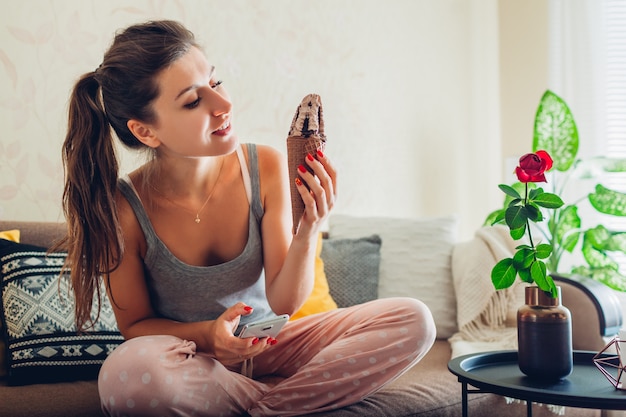 Young woman eating chocolate ice-cream in cone sitting on couch at home and using smartphone.