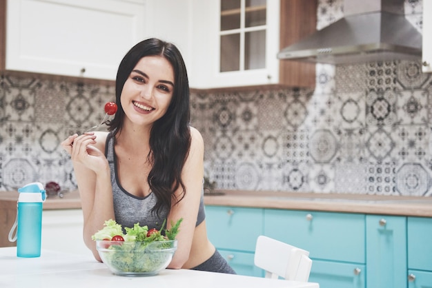 Молодая женщина ест полезный салат с помидорами черри на кухне после фитнес-сессии