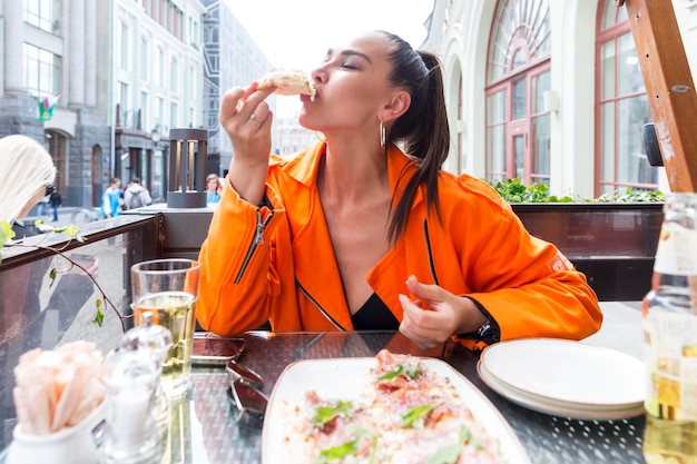 通りの街のカフェでピザを食べる若い女性 明るいオレンジ色のジャケットを着た美しいブルネットがおいしい一口を取る