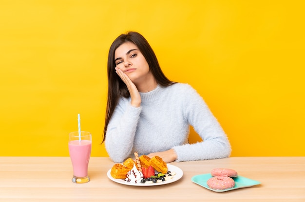 孤立した黄色の不幸と欲求不満の上にテーブルでワッフルとミルクセーキを食べる若い女性