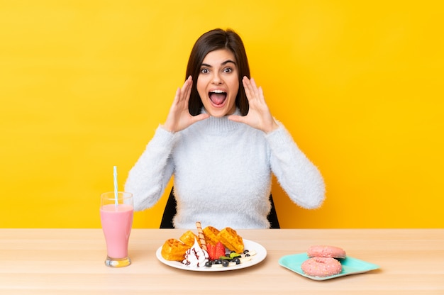 Young woman eating waffles and milkshake in a table over yellow wall shouting with mouth wide open