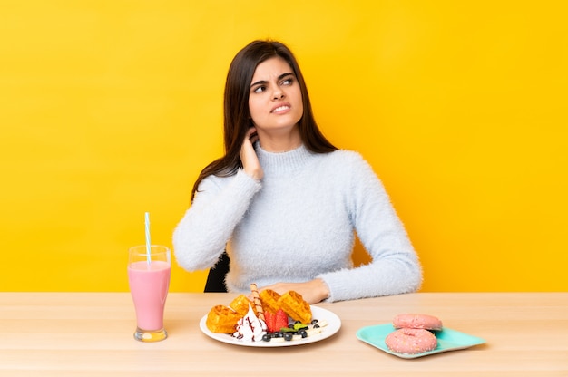 Young woman eating waffles and milkshake in a table over yellow wall thinking an idea