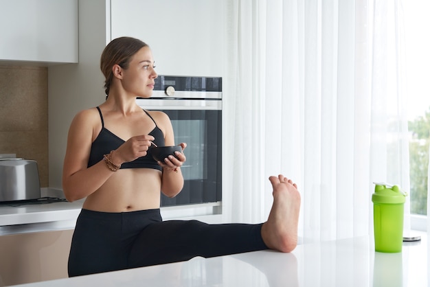 Young woman eating while stretching in kitchen with leg on table