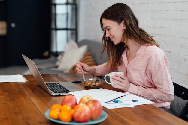 Young woman eats breakfast sitting in front of her laptop.