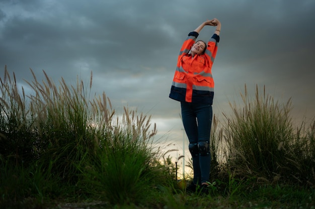 Young woman in engineer uniform and high visibility with raised arms standing on grassy field at sunset The concept of relax time after work