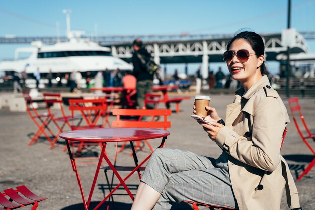 米国サンフランシスコの港近くの屋外カフェに座ってコーヒーを楽しむ若い女性。背景に青い空とオークランド ベイ ブリッジ。携帯電話を持って明るく微笑むアジアの女性。