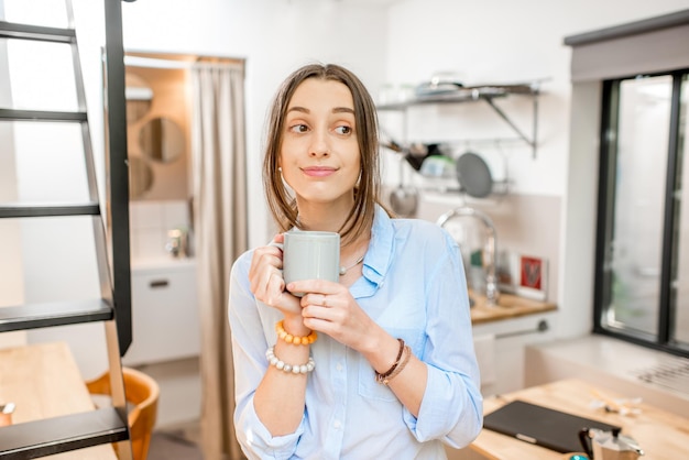 カントリーハウスの小さなキッチンに立ってコーヒーを楽しんでいる若い女性