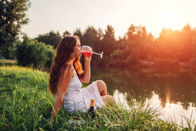 Giovane donna che gode del bicchiere di vino sulla sponda del fiume al tramonto.