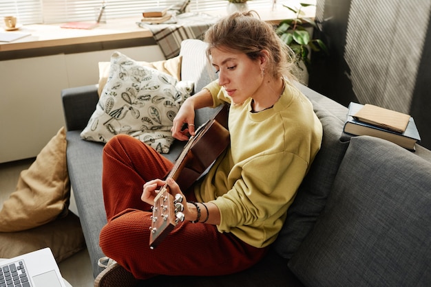 Foto giovane donna che si diverte a suonare la chitarra sul divano nel soggiorno di casa