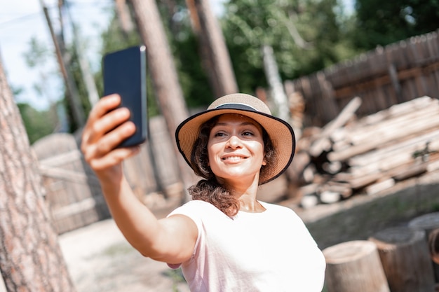 밀짚모자를 쓰고 여름 햇살을 즐기는 젊은 여성은 전화 카메라로 자신의 사진을 찍고, 전화로 셀카를 찍고, 나무가 있는 시골 마당의 배경입니다.