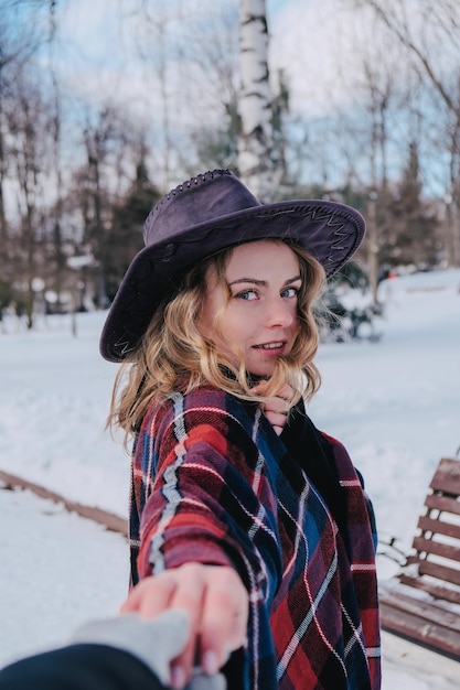 Young woman enjoying winter weather in the snow park. Cold weather. Winter fashion, holidays, rest, travel concept. Cowboy hat and poncho. Curly blond girl