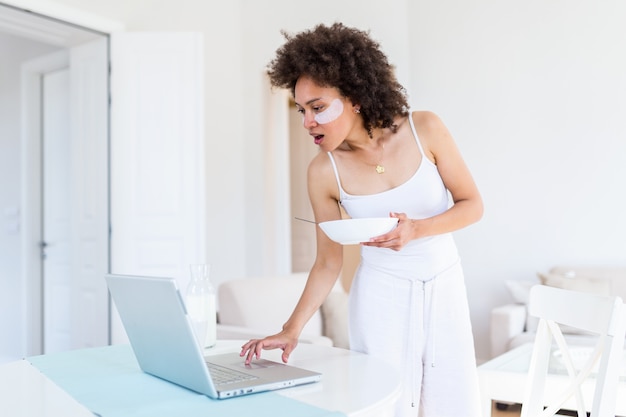 young woman enjoys eating corn flakes for breakfast while working on her laptop in the morning.