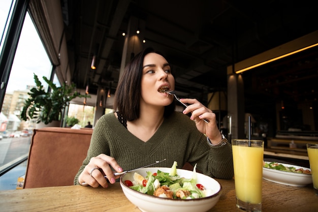 若い女性はおいしい食事を楽しんでいます。健康的なシーザーサラダをゆっくりと食べる茶色の髪の魅力的な女性