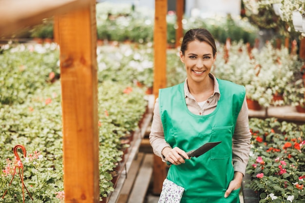 Young woman entrepreneur standing in front of tables of potted flowers and smiling at the camera. She is wearing a green apron and working in plant nursery or greenhouse.