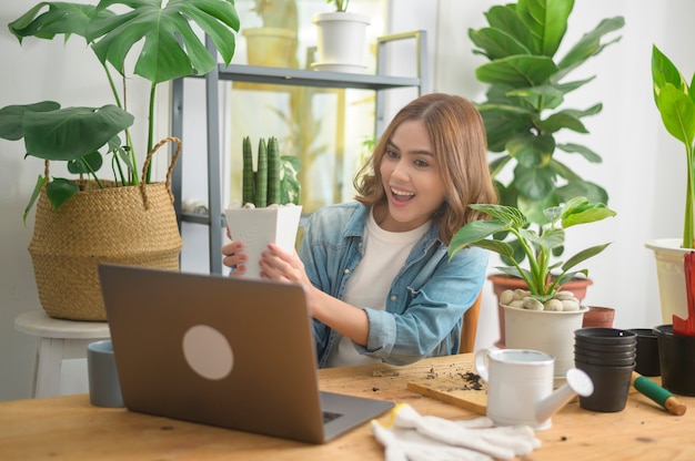 A young woman entrepreneur working with laptop presents houseplants during online live stream at home