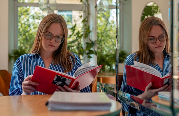 Молодая женщина в очках читает книгу в современном кафе с растениями и читателем дневного света в кофейне
