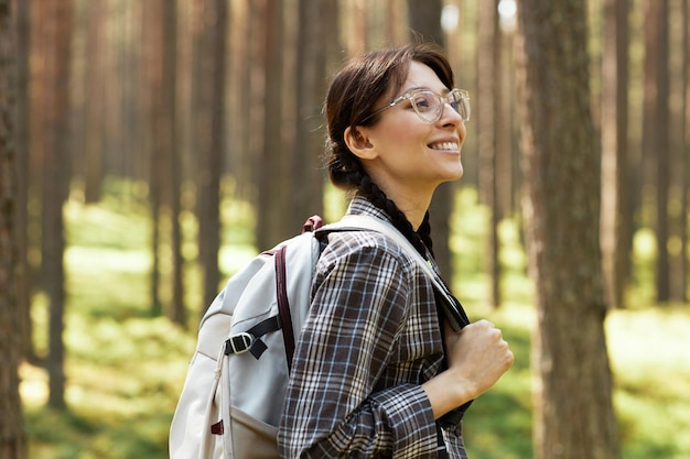 Молодая женщина в очках с рюкзаком за спиной наслаждается природой, гуляя по лесу