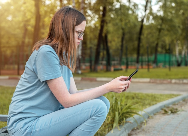 Young woman in eyewear sitting on bench alone and using mobile phone person looking on smartphone in...