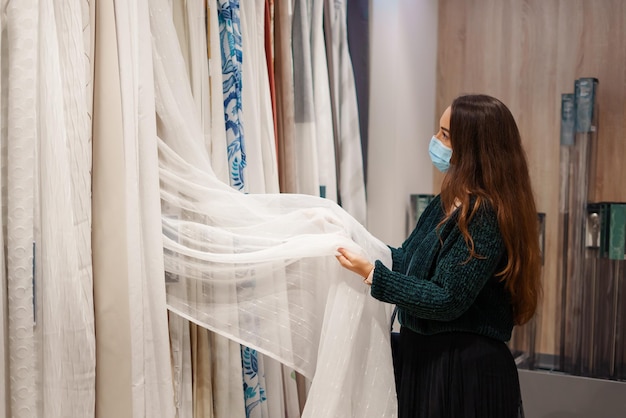 Young woman in face mask choosing tulle and curtains in fabric store doing shopping during covid