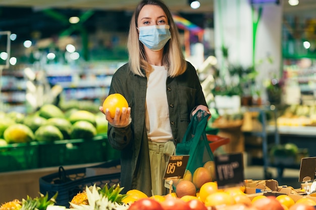 La giovane donna in maschera protettiva medica in faccia sceglie e raccoglie in eco-bag verdure o frutta al supermercato la donna in piedi in un mercato di alimentari vicino al bancone acquista in un pacchetto riutilizzabile