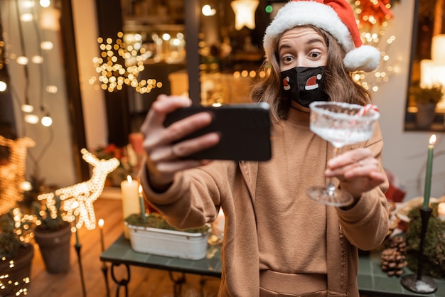 Giovane donna in maschera facciale che celebra da sola le vacanze di capodanno a casa, facendo una videochiamata al telefono con gli amici. concetto di quarantena e autoisolamento durante l'epidemia nei giorni festivi