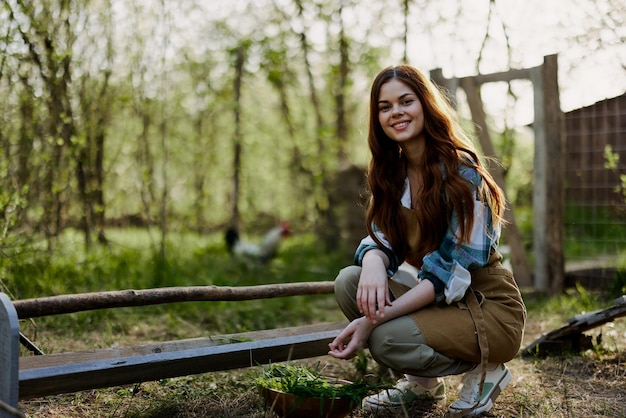 A young woman farmer sits at a bird feeder and checks the composition of the grain to feed the chickens in a pen in the countryside